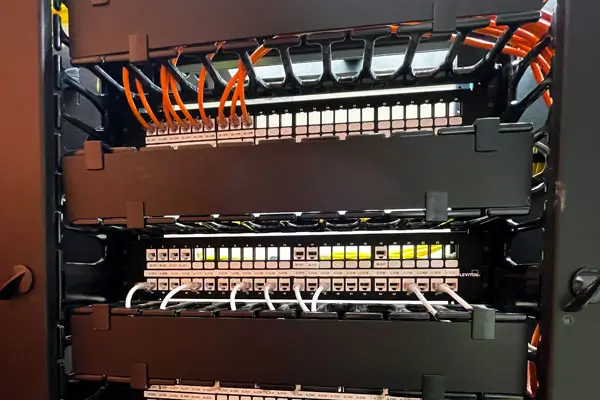 A network server rack with multiple rows of organized cat six network cables and connectors. Orange cables are neatly arranged, connecting various ports on the panels within a dark metal frame.