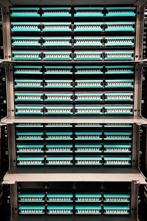 A server rack filled with rows of turquoise fiber optic bulkhead connectors, organized neatly in a secure data center environment. The equipment is arranged horizontally across multiple shelves.