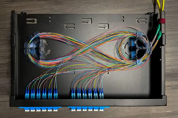 An organized fiber optic cable management tray with colorful wires arranged in a symmetrical, figure-eight pattern. The cables are neatly connected to blue adapters on one side, displaying meticulous fiber optic infrastructure.