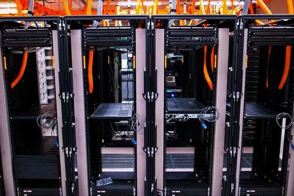 A row of empty black server racks in a data center, with orange cable housing running across the top. The setting is well-lit, showcasing the organized methods of decommissioning used to remove and dispose of technological components.